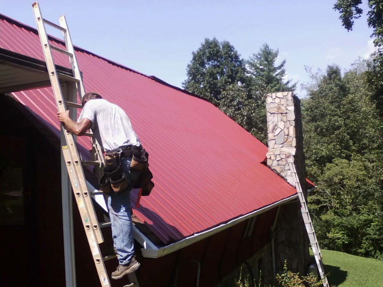 Roofer on a ladder beside a red metal roof and stone chimney — Soffit & Fascia Repair by Roofing Brothers in Keithsburg